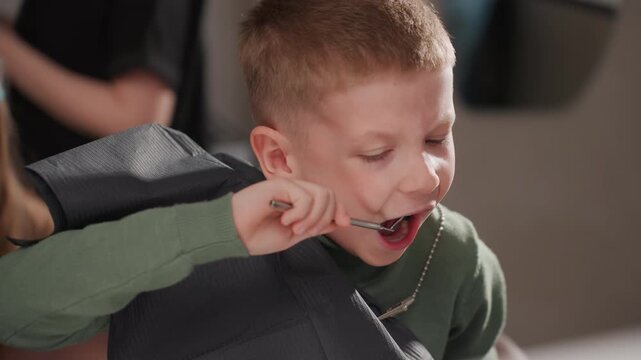 Young boy sits in dental chair wearing protective bib while holding mirror accompanied by woman with face shield. Dental office setting visible including medical equipment and countertop