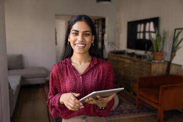 Confident professional Indian woman stands in modern living room, holding tablet, smiling at camera, portraying realtor, designer or entrepreneur working with clients in real estate or interior design © fizkes