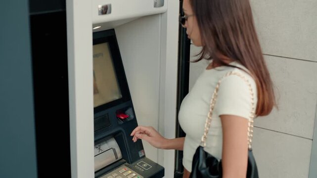Young brunette woman in sunglasses using an automated teller machine, inserting her credit card to withdraw cash and complete her financial transaction at an outdoor bank terminal