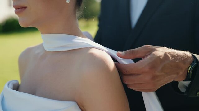 Groom in a black tuxedo tenderly adjusts the white fabric detail on his bride's dress, showcasing a moment of care and intimacy during their outdoor wedding celebration
