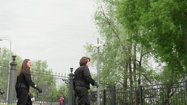 Close up of three people in black padded outfits walking along wrought iron fence with green trees and overcast sky. group includes two women one man all wearing protective gear
