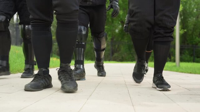 Close up of multiple people walking in unison wearing black clothing and knee pads on paved path with green grass and trees in background. synchronized movement emphasizes coordinated