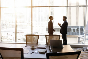 Naklejka premium Businessmen talking in office boardroom, discuss project strategy, planning business decisions standing next to window overlooking city. Corporate communication, professional negotiation, partnership
