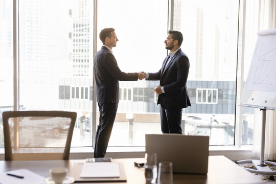 Two multiethnic businessmen shaking hands, concluding agreement in modern corporate office meeting room. Completing business deal with handshake, starting partnership, successful negotiation outcome