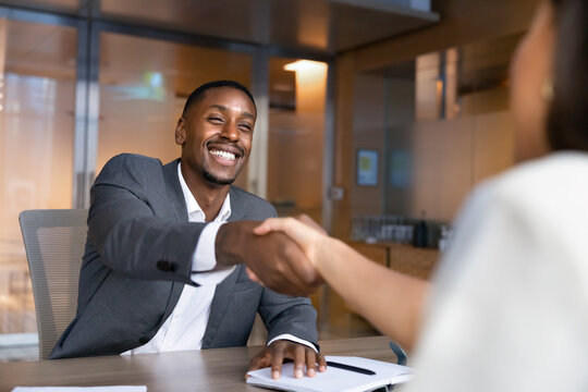 African businessman sitting at office desk, smiling, shaking hands with business colleague or partner during corporate meeting. Recruitment, partnership agreement and successful negotiations concept