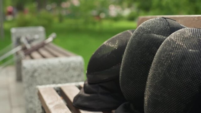 Close up of person in black fencing gear reaching for helmets on outdoor bench. Fencing equipment including masks and gloves visible with green park background. Soft natural light highlights