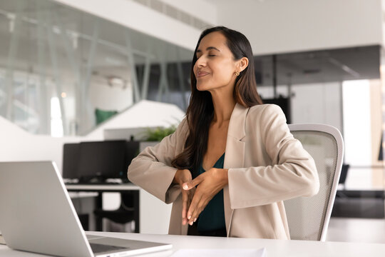 Businesswoman sits at office desk with folded palms, take break, making calming breathing exercise, practicing mindfulness meditation for stress management, fatigue relief. Relaxation at workplace