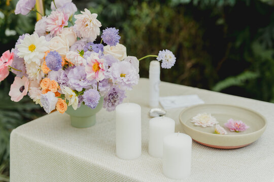 Elegant floral arrangement with soft purple and white flowers displayed beside white candles and tableware in a refined wedding or event setting.