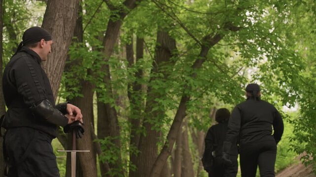 Close up of two men in black medieval style clothing standing among trees one holding sword. third men joins them as they walk away through forested area with dense green foliage and tall