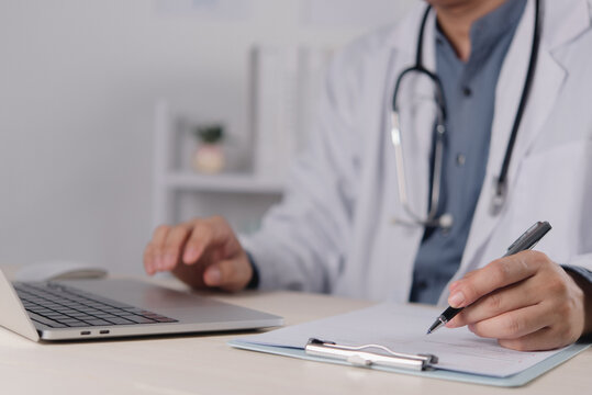 Healthcare professional writing on clipboard while using laptop for clinical documentation and digital medical office workflow workspace