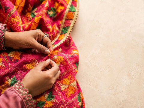 Phulkari embroidery artisan hand stitches vivid floral patterns on a bright punjabi dupatta using colorful thread and a fine needle while traditional textile details stand out on a pale background