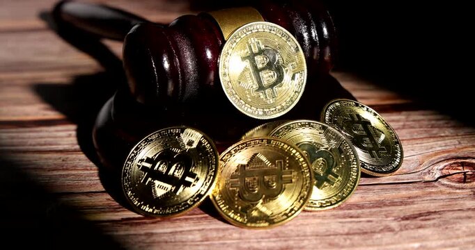 Horizontal rotating tracking camera pass around an isolated wooden judge gavel and sounding block positioned next to glowing golden bitcoin cryptocurrency coins on dark backdrop.