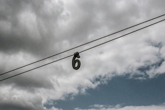 Metallic number six hangs from wires against cloudy sky background. A singular numeral suspended mid-air, suggesting a countdown or a specific point.