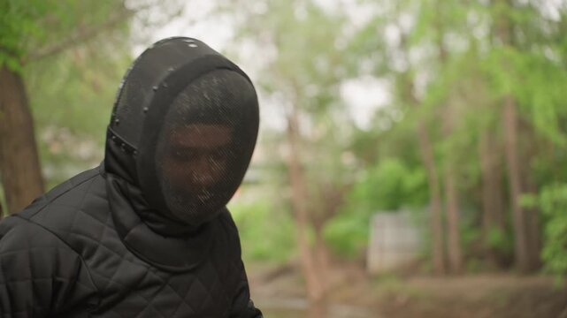 Close up of person in black fencing gear practicing sword techniques outdoors. person wearing protective mesh helmet and padded jacket demonstrates various stances with two swords