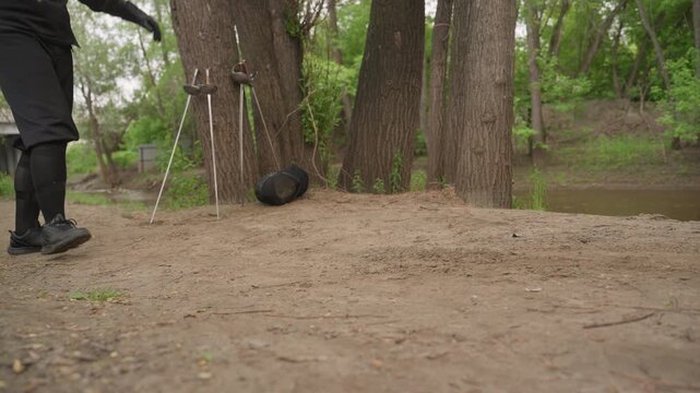 Close up of two people practicing sword fighting outdoors on dirt path surrounded by trees. One person holds fencing equipment while other moves into position with swords and protective gear