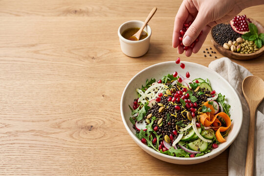 Hand adding pomegranate seeds to a fresh lentil salad with vegetables in a bowl on a wooden table