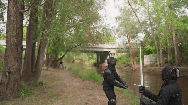 Close up of multiple people dressed in black protective gear practicing sword fighting along riverside path with trees and overpass. participants wield long swords demonstrating various