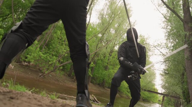 Two men in black protective gear engage with swords along riverbank surrounded by green trees and overcast sky. One man holds his sword high while other prepares to strike both wearing