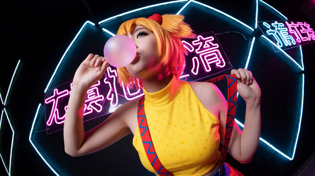 Vibrant young woman wearing yellow cosplay costume and red suspenders blows a large pink bubble gum sphere in front of glowing neon signs at night