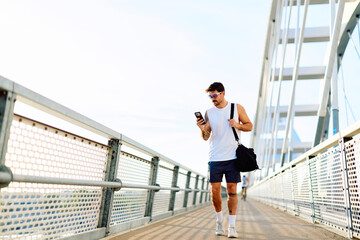 Naklejka premium Man carrying a gym bag and checking his smartphone while walking on a modern bridge after a fitness workout