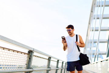 Naklejka premium Young man wearing sportswear and sunglasses walking on a modern bridge, using his smartphone during a morning workout break