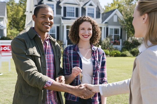 Real estate milestone scene of diverse couple receiving keys to new home with agent and sold sign