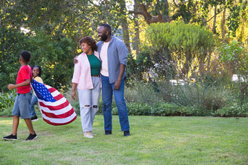 Obraz premium African American family walking across grassy lawn, carrying American flag near flowerbed