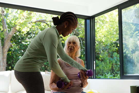 African American trainer wearing green top guiding Senior woman holding purple dumbbells at home