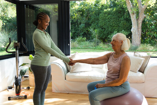 Two women, senior sitting on gym ball African-American trainer holding hands aiding balance at home