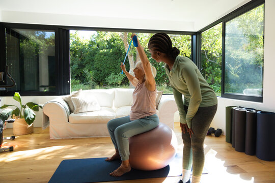Senior woman sitting on exercise ball holding blue band, African American coach guiding at home