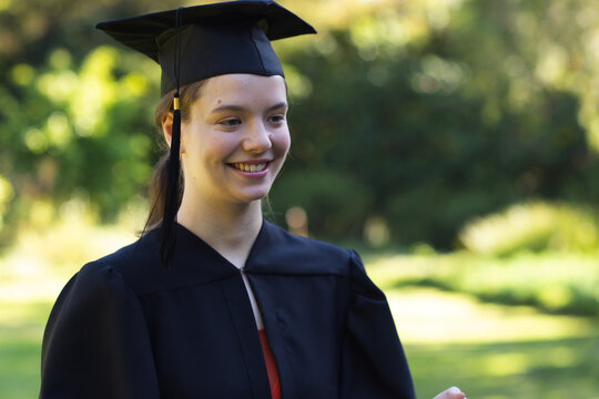 Woman wearing graduation cap and black gown smiling while standing on sunlit lawn