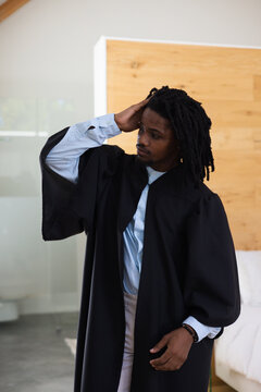 African American man wearing academic gown adjusting dreadlocks in bedroom by frosted glass ensuite