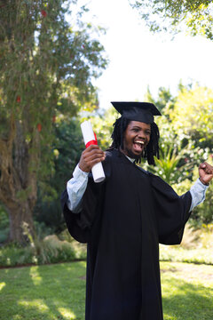 African man laughing while holding red ribbon diploma and wearing mortarboard on sunlit lawn