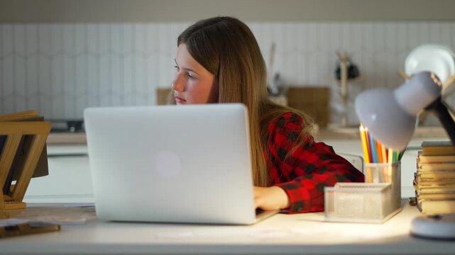 Teen girl studying on laptop at home. Student typing on computer at desk. Girl doing homework with book stack nearby. Student learning online at home desk. Teen studying with lamp light in kitchen.