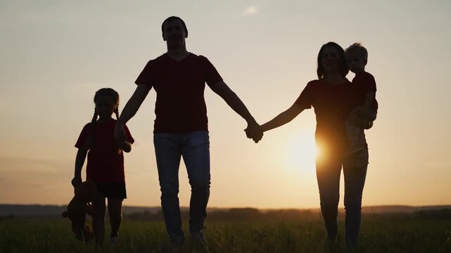 Happy family walks across grassy field at sunset. Parents hold hands with children in golden light. Mother carries little son. Daughter holds teddy bear. Family silhouettes enjoy evening walk.