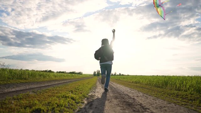 Young girl runs with kite. Child enjoys play during summer. Silhouette of girl moving fast. Colorful kite flies over green field. Run down dirt path at sunset. Nature surrounds wide green field.