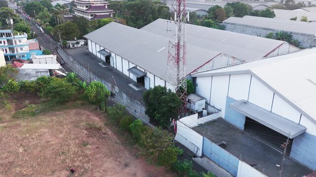 Aerial drone view of a dense industrial area featuring large warehouse buildings, factory rooftops, surrounding roads, vehicles, trees, and urban infrastructure. The composition highlights the contras