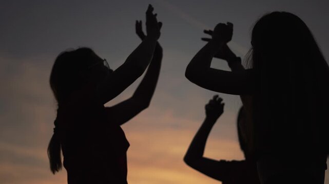 Silhouette of friends dancing at sunset. Women raise hands against orange sky. Group of people dance together at dusk. Dark silhouette figures celebrate outdoors. Friends enjoy sunset party dancing.