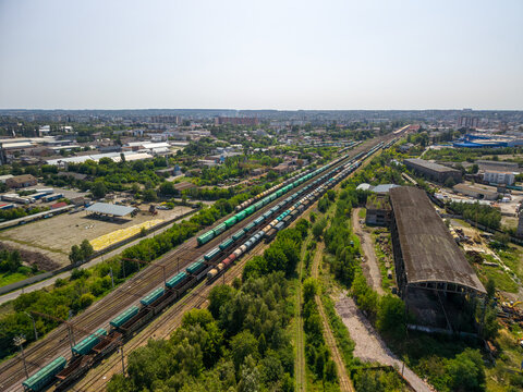 High angle aerial shot captures long cargo trains on parallel tracks next to old industrial warehouse building, green vegetation, and distant city buildings under clear sunny sky