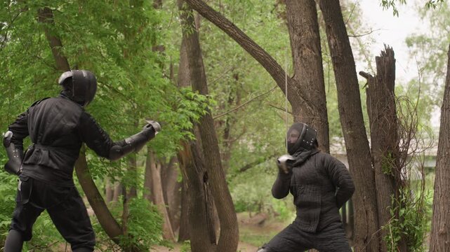 Two people in black protective armor engage in sword fight within dense forest setting demonstrating dynamic movements and techniques. scene features close up views of their stances weapon