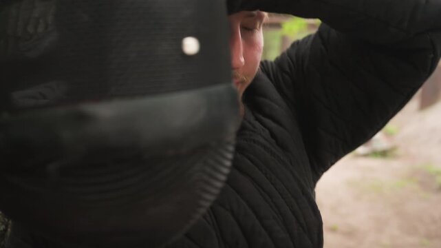 Close up of man in black quilted clothing holding and putting on medieval style armor pieces outdoors. man interacts with helmet showing details like mesh interior against earthy background