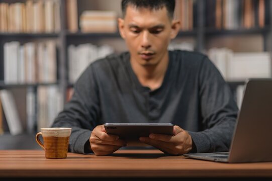 Man Using Tablet at Desk with Bookshelf Background and Laptop, Focused on Screen