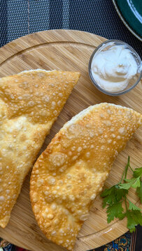 Traditional crispy chebureki, deep-fried meat-filled pastries served with sour cream and fresh parsley on a wooden board.