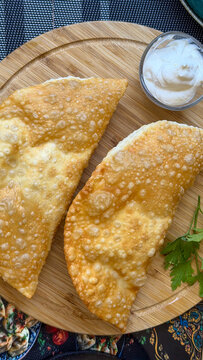 Traditional crispy chebureki, deep-fried meat-filled pastries served with sour cream and fresh parsley on a wooden board.