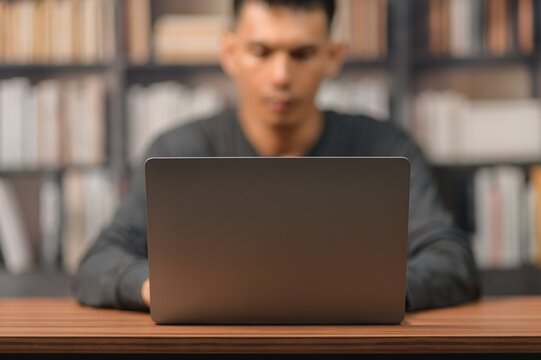 Young Man Working on Laptop in Library with Bookshelf in Background