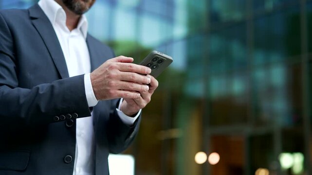 Close up of mature businessman hands using a smartphone while wearing a formal suit. Male professional browsing mobile content on phone with a blurred urban background near a modern office building.