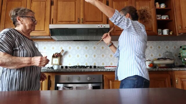 Joyful daughter dancing and singing with a wooden spoon for her elderly mother in the kitchen. Happy family moment representing intergenerational bonding, fun, and positive domestic life