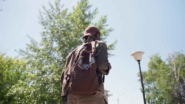 Close up of man wearing outdoor gear with backpack walking away from camera in bright daylight. Brown jacket and khaki pants visible under large brown hiking bag on his back. Outdoor scene