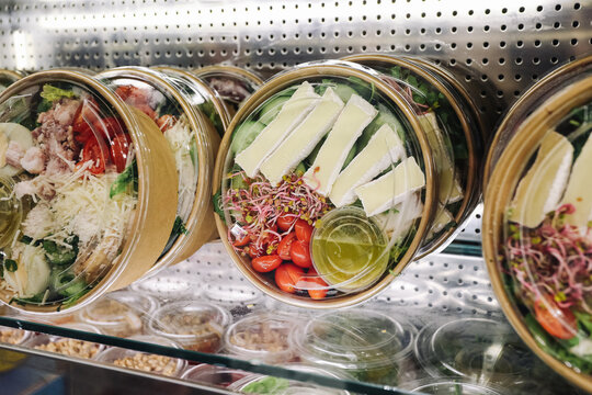 Fresh takeaway salads in round containers on refrigerated store shelf. Vegetables cheese sprouts and dressing form ready meals. Healthy convenience food background. Retail deli selection.