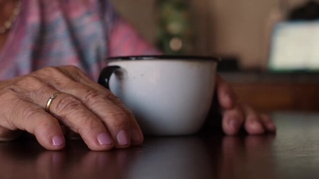 Close-up view of a senior woman's wrinkled hands with a wedding ring and manicured nails, nervously tapping her fingers on a dark wooden table next to a white enamel mug